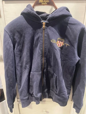 Marinblå hoodie från GANT - Mörkblå hoodie från GANT med dragkedja framtill och broderad logga samt sköld på bröstet. Tröjan har huva, ribbade muddar och två stora fickor framtill. Perfekt för en avslappnad och sportig stil. Materialet känns mjukt och bekvämt.