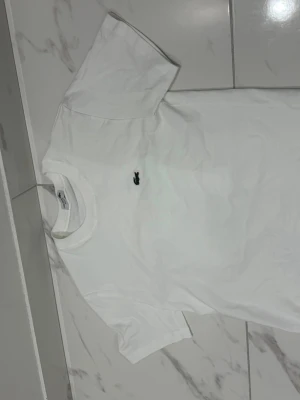 Vit t-shirt från Lacoste - Klassisk vit t-shirt från Lacoste med den ikoniska lilla krokodilloggan broderad på bröstet. T-shirten har rund hals och korta ärmar, perfekt till alla tillfällen. Tillverkad i mjuk bomull för skön känsla.