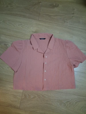 Rosa kortärmad blus med krage - Trendig rosa blus från Zalora med korta puffärmar och klassisk krage. Blusen har en lätt skrynklig struktur och knäpps framtill med vita knappar. Perfekt för en avslappnad men stilsäker look.