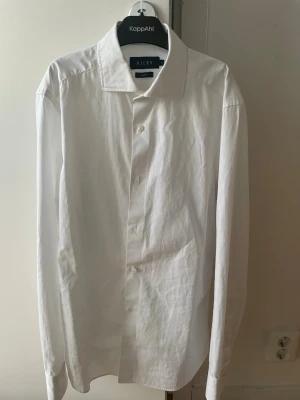 Vit klassisk skjorta från Riley - Säljer en stilren vit skjorta från Riley med klassisk krage och knäppning framtill. Skjortan har lång ärm och är tillverkad i bomull, vilket gör den både bekväm och lätt att matcha till olika outfits. Slim fit.