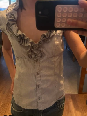 Randig blus med volang och knappar - Säljer en söt blus med smala grå och vita ränder, djup v-ringning och volangdetaljer längs halsen. Blusen har korta puffärmar och knappar framtill. Perfekt för dig som gillar feminina detaljer och en figurnära passform.