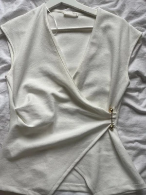 Vit omlottopp från Zara - Snygg vit omlottopp från Zara med v-ringning och guldfärgade knappdetaljer i sidan. Toppen är ärmlös och har en elegant, figurnära passform. Perfekt att styla med jeans eller kjol för en trendig look.