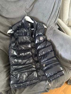 Svart glansig dunväst från Moncler - Snygg svart dunväst från Moncler med glansig finish och hög krage. Västen har två dragkedjefickor framtill och en Moncler-logga på bröstet. Perfekt för lager på lager och ger en cool streetwear-look.