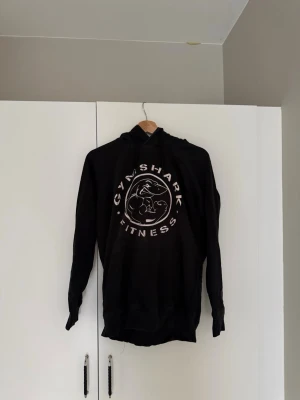 Svart Gymshark hoodie med tryck - Säljer en svart hoodie från Gymshark med stort vitt tryck på bröstet där det står 'GYMSHARK FITNESS' och en haj-logga. Hoodien har huva och ribbade muddar vid ärmar och nederkant. Perfekt för gymmet eller chill dagar. Bra skick. Storlek S.
