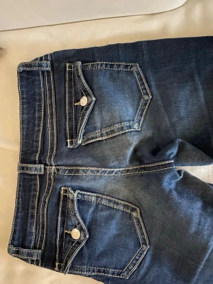 Mörkblå bootcut jeans med kontrastsömmar - Snygga mörkblå bootcut jeans med vita kontrastsömmar och dekorativa ficklock bak. Klassisk femficksmodell med knappdetaljer och lätt utsvängda ben. Perfekta för en avslappnad och trendig look.