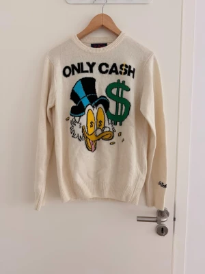 MC2 Saint Barth Scrooge McDuck Disney knit sweater size S - Elevate your knitwear collection with this unique MC2 Saint Barth sweater featuring Disney's Scrooge McDuck! Made from a luxurious blend of wool and cashmere, this off-white sweater is incredibly soft and comfortable. The iconic "ONLY CASH" design, Scrooge McDuck graphic, and embroidered cuff logo make it a true statement piece. In excellent condition, never used, size S. Don't miss out on this playful and stylish find!