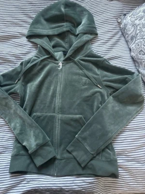 Grön velourhoodie från Juicy Couture - Mjuk och skön grön hoodie i velour från Juicy Couture. Dragkedja framtill, fickor och broderad JC-logga på bröstet. Klassisk huva med dragsko och ribbade muddar vid ärmar och nederkant. Perfekt för en chill och trendig look.