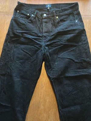 Svarta manchesterbyxor från H&M - Säljer ett par svarta manchesterbyxor från H&M med klassisk femficksmodell och raka ben. Byxorna har synliga nitar och knappdetaljer framtill samt breda manchesterränder för en retrovibe. Perfekta för dig som gillar en avslappnad men ändå stilren look.