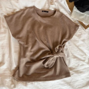 Beige topp med knyt från Zara - Supersnygg beige topp från Zara med rund halsringning och korta ärmar. Toppen har en unik knytdetalj i midjan som ger en cool och trendig look. Perfekt att styla med jeans eller kjol för en avslappnad men chic outfit.