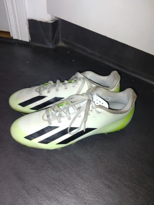 Adidas vita och lime fotbollsskor - Säljer ett par snygga fotbollsskor från Adidas med vit ovandel, limegröna detaljer och klassiska svarta stripes på sidan. Skorna har snörning och är tillverkade i syntetmaterial som ger bra grepp och komfort på planen.