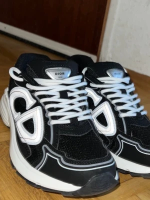 Svarta Dior sneakers med vita detaljer - Säljer ett par svarta Dior sneakers med vita detaljer och stora CD-loggor på sidorna. Skorna har vita snören, meshpaneler för extra andningsförmåga och en chunky vit sula.