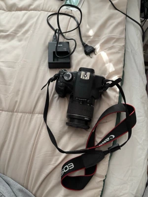 Canon EOS 1200D systemkamera - Canon EOS 1200D systemkamera med svart kamerahus och ett Canon Zoom Lens EF-S 18-55mm objektiv. Kameran har en tydlig display, flera inställningsrattar och medföljande axelrem med röd kant. Batteriladdare och strömkabel ingår.