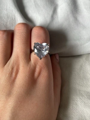 Hjärt ring  - En silvrig hjärt ring med en diamant i mitten som är format som ett hjärta. Diamanten är lila liknande men främst genomskinlig. Perfekt till dig som vill ha till en liten tutch.