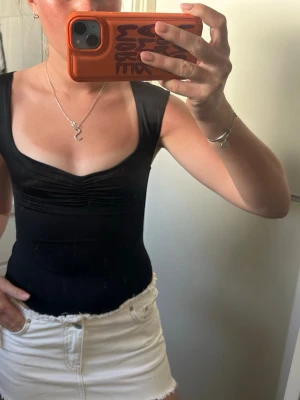 Svart body med djup urringning - Säljer en svart body med djup urringning och rynkdetalj vid bysten. Bodyn har breda axelband och är tillverkad i ett stretchigt, mjukt material som sitter tight mot kroppen. Perfekt att styla med jeans eller kjol för en trendig look.