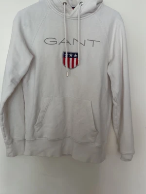 Vit hoodie från GANT med logga - Snygg vit hoodie från GANT med stor broderad logga och amerikanskinspirerat sköldmärke på bröstet. Hoodien har huva med dragsko och en stor magficka. Tillverkad i mjukt material, perfekt för en avslappnad och trendig look.