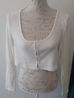 Vit croppad ribbad topp med knappar - Trendig vit croppad topp med långa ärmar och ribbad struktur. Toppen har djup rundringning och knappar framtill för en snygg detalj. 