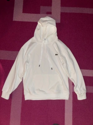 Vit hoodie från Lacoste med huva - Säljer en clean vit hoodie från Lacoste med klassisk krokodil-logga på bröstet. Hoodien har dragsko i huvan och en stor magficka. Tillverkad i mjukt material som känns skönt mot huden. Perfekt för dig som gillar stilrena plagg med ikonisk branding.