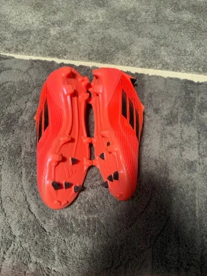 Röda Adidas fotbollsskor utan snörning - Säljer ett par röda Adidas fotbollsskor med svarta ränder på sidorna. Skorna har en modern, strömlinjeformad design utan snörning och är tillverkade i syntetmaterial för bra passform och grepp. Perfekta för dig som vill ha stilrena skor, 