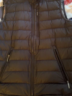 Svart dunväst med dragkedjor - Säljer en svart dunväst med quiltad design och två snedställda fickor med dragkedja. Västen har hög krage och stängs med dragkedja framtill. Perfekt för lager-på-lager och riktigt skön att bära under kyliga dagar.