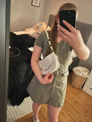 Grön kortärmad skjortklänning - Snygg grön skjortklänning med v-ringning, korta ärmar och knappar hela vägen framtill. Klänningen har fyra fickor och är tillverkad i ett lätt bomullstyg, perfekt för varma dagar. Enkel och stilren design som passar till många tillfällen. Storlek 170 men passar dam/kvinna och flicka i XS, S och liten M. 