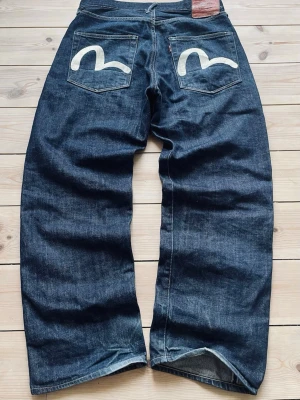 Evisu jeans med målad logga bak - Säljer ett par mörkblå Evisu jeans med bred passform och ikoniska handmålade vita måsloggor på bakfickorna. Klassisk femficksmodell i kraftig denim med läderpatch i midjan. Perfekta för dig som gillar streetwear och vill sticka ut.