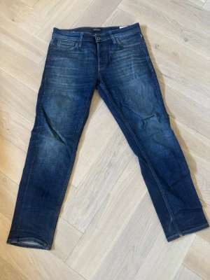 Mörkblå Tapered/Mike jeans från Jack&Jones - Säljer ett par mörkblå jeans med klassisk femficksdesign och snygga slitningar på låren. Jeansen har raka ben och är tillverkade i bomull med stretch för extra komfort. Perfekt för dig som gillar en modern och avslappnad look.