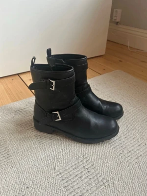 Svarta boots med spännen - Snygga svarta boots i skinnimitation med två silverfärgade spännen och grov sula. Klassisk modell med rund tå och dragflikar upptill. Perfekta för en edgy look till jeans eller kjol.