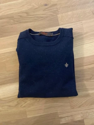 Mörkblå stickad tröja Morris - Snygg mörkblå stickad tröja från Morris med rund halsringning och diskret broderad logga på bröstet. Tröjan har långa ärmar och ribbade muddar vid ärmslut och nederkant. Perfekt för lager på lager och stilrena outfits. Skriv vid funderingar!