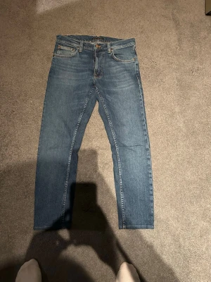 Blå jeans från Nudie Jeans - Säljer ett par klassiska blå jeans från Nudie Jeans med raka ben och fem fickor. Jeansen har en snygg tvätt och detaljerade sömmar på bakfickorna. Tillverkade i slitstark denim med en tidlös look.