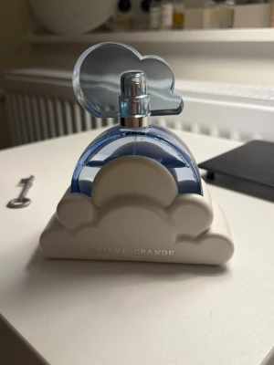 Ariana Grande Cloud parfym 100ml - Unik parfymflaska från Ariana Grande formad som ett blått moln med vit molnbas. Flaskan har en lekfull design med transparent blå plast och vit plastdetalj. Perfekt för dig som gillar snygga och kreativa parfymflaskor.