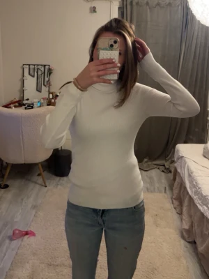 Vit ribbad polotröja - Säljer en vit, ribbstickad polotröja med lång ärm. Tröjan har en tight passform och hög krage, perfekt för lager på lager. Materialet ser ut att vara mjukt och stretchigt, vilket gör den bekväm och enkel att styla till olika outfits.