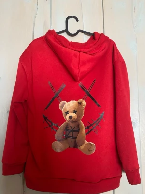 Röd hoodie med nalletryck - Cool röd hoodie med stor ficka framtill och huva med dragsko. På ryggen finns ett tryck av en nallebjörn med svarta detaljer och korsade streck, vilket ger en edgy vibe. Hoodien är gjord i mjukt material, perfekt för en avslappnad streetwear-look.