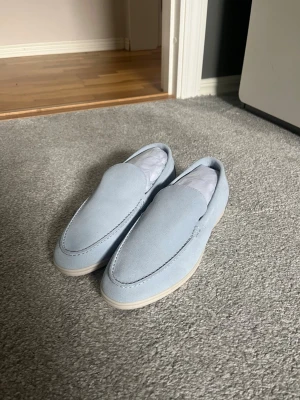 Ljusblå loafers från Loro Piana - Säljer ett par stilrena ljusblå loafers från Loro Piana, modell Summer Walk. Skorna är tillverkade i mjukt mockaskinn och har en klassisk, minimalistisk design med rundad tå och diskreta sömmar. Perfekta för en avslappnad men ändå elegant look.