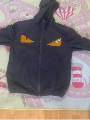 Svart hoodie med gula ögon - Cool svart hoodie med dragkedja och huva. På bröstet finns två gula ögon i glansigt material som ger ett unikt och edgy uttryck. Perfekt för dig som vill sticka ut i mängden. Hoodien har fickor på sidorna och är tillverkad i ett mjukt tyg.