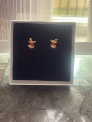 Juliga örhängen med röda kulor - Säljer ett par örhängen formade som små röda julkulor med guldfärgade detaljer och blad upptill. Kulorna har en rad glittrande stenar som ger extra bling. Perfekta för dig som vill ha en festlig touch till din look.