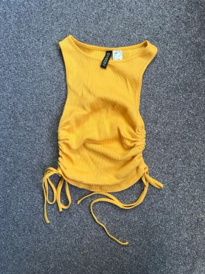 Orange ribbad linnetopp från H&M Divided - Snygg gul ribbad linnetopp från H&M Divided med trendiga snörningsdetaljer i sidorna. Toppen är ärmlös och har en croppad passform som passar perfekt till höga jeans eller kjol. Materialet är mjukt och stretchigt för en bekväm känsla.