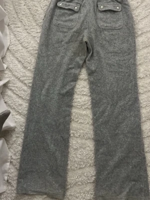 Grå mjukisbyxor Juicy Couture - Mysiga grå mjukisbyxor från Juicy Couture i mjukt velourmaterial. Byxorna har raka ben, två bakfickor med lock och knappar samt en snygg broderad JC-logga. Perfekta för chill dagar eller när du vill vara extra bekväm.