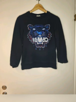 Svart sweatshirt från Kenzo med tiger - Säljer en svart sweatshirt från Kenzo med det ikoniska tigerhuvudet broderat i blått, vitt och lila på bröstet samt texten 'KENZO PARIS'. Tröjan har rund halsringning, långa ärmar och är gjord i mjukt bomullsmaterial. Perfekt för dig som gillar streetwear och vill sticka ut.