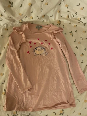 Rosa långärmad nattlinne Alfons Åberg barn st 122/128 - Söt rosa långärmad nattlinne från Alfons Åberg med volangdetaljer på axlarna. Tryck framtill med ett ansikte och rosa hjärtan. Tillverkad i mjuk bomull. Barn storlek 122/128