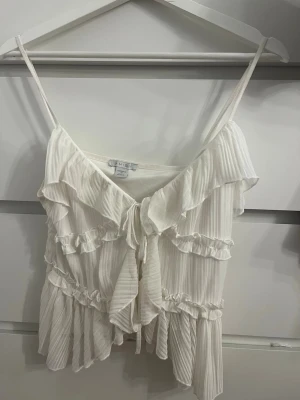 Vit volangtop från MISU med knyt - Supersöt vit topp från MISU med tunna axelband, volanger och knyt framtill. Toppen har en luftig och romantisk känsla med flera lager volanger och lätt ribbad struktur. Perfekt för dig som gillar en feminin och trendig stil.