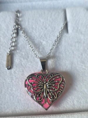 Halsband med hjärta och fjäril som lyser lila  - Snyggt halsband i silverfärgad metall med ett hjärtformat hänge. Hänget har en detaljrik fjäril i metall ovanpå en rosa bakgrund som lyser lila i mörker efter att ha absorberat dagsljus eller solljus. Solljus ger snabbast resultat. Vänd på hänget en stund för bästa effekt. Kedjan är tunn och justerbar i rostfritt stål. Både kedjan & hänget tål vatten utan att missfärgas. Perfekt för dig som gillar unika smycken.