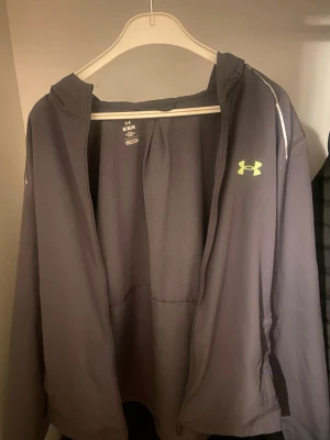 Grå hoodie från Under Armour - Snygg grå hoodie från Under Armour med dragkedja och huva. Har ett gult logotryck på bröstet och vita detaljer längs axlarna. Tillverkad i ett lätt och ventilerande material som passar perfekt till träning eller chill. Praktiska fickor framtill.