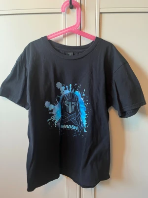 Svart Smash T-shirt från smashintopieces - Cool svart t-shirt från smash into pieces med ett blått och vitt Smash-motiv på bröstet. T-shirten har rund hals, korta ärmar och är gjord i mjuk bomull. Äkta merch från smashintopieces 