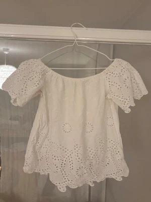 Vit broderad blus med puffärm - Söt vit blus med broderade hålmönster och volangkant. Blusen har puffiga korta ärmar och en vid, luftig passform. Off shoulder-modell med elastisk halsringning. Tillverkad i bomullstyg som känns lätt och somrig.