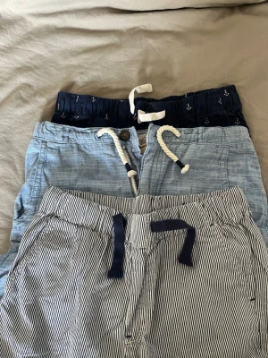 Randiga och ljusblå shorts med snörning - Tre par shorts i olika stilar: ett par randiga i vitt och marinblått med mörkblå snörning, ett par ljusblå i tunn bomull med vit snörning och ett par mörkblå med ankarmönster och vit snörning. Alla har avslappnad passform och elastisk midja för skön känsla.