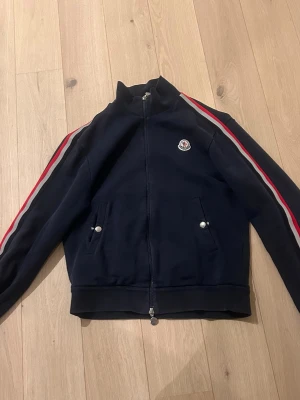 Marinblå ziptröja från Moncler - Marinblå ziptröja från Moncler med röda och vita ränder längs ärmarna. Tröjan har hög krage, dragkedja framtill och två fickor med tryckknappar. Moncler-logga på bröstet. Tillverkad i mjukt material, perfekt för en aktiv stil. Storlek M. Hör av er vid fler bilder! Pris kan diskuteras.