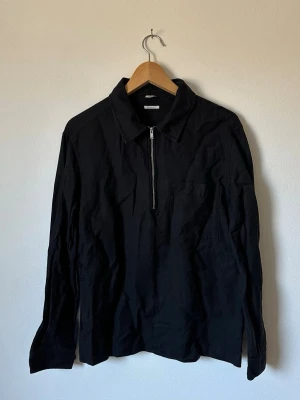 Svart Arket overshirt med dragkedja - Stilren svart Arket overshirt med krage och halv dragkedja framtill. Långärmad modell med knapp vid ärmslut och bröstficka. Perfekt att bära över en t-shirt för en avslappnad look. Materialet ser ut att vara bomull eller bomullsblandning.