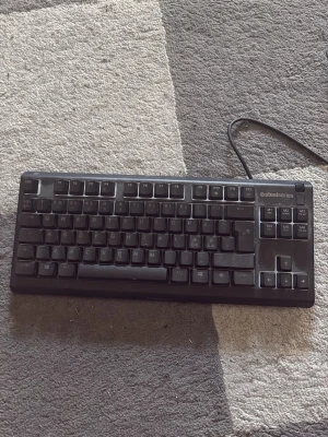 SteelSeries Apex 3 TKL tangentbord - SteelSeries Apex 3 TKL är ett kompakt, svart tangentbord med nordisk layout och tyst membranteknik. Perfekt för gaming eller studier, med programmerbara knappar och robust plastkonstruktion. Ansluts via USB-kabel.