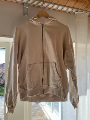 Beige zip hoodie från vailent - En enkel och clean beige hoodie med dragkedja framtill och två stora fickor. Hoodien har huva och ribbade muddar vid ärmar och nederkant. Perfekt för en avslappnad och chill stil. Materialet känns mjukt och bekvämt, troligtvis bomull eller bomullsblandning.