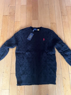 Svart kabelstickad tröja Polo Ralph Lauren - Kabelstickad svart tröja från Polo Ralph Lauren med klassisk röd logga broderad på bröstet. Tröjan har rund halsringning och ribbade muddar vid ärmslut och nederkant. Perfekt för en stilren och tidlös look.